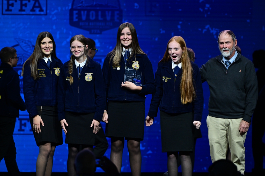 Carroll County Middle FFA alumnae Magen Key (second from left), Natalie Culler (center) andMadelyn Caviness (second from right) celebrate on stage with 2022-23 National FFA Secretary Jessica Herr (far left) and their advisor, John Carpenter (far right).
