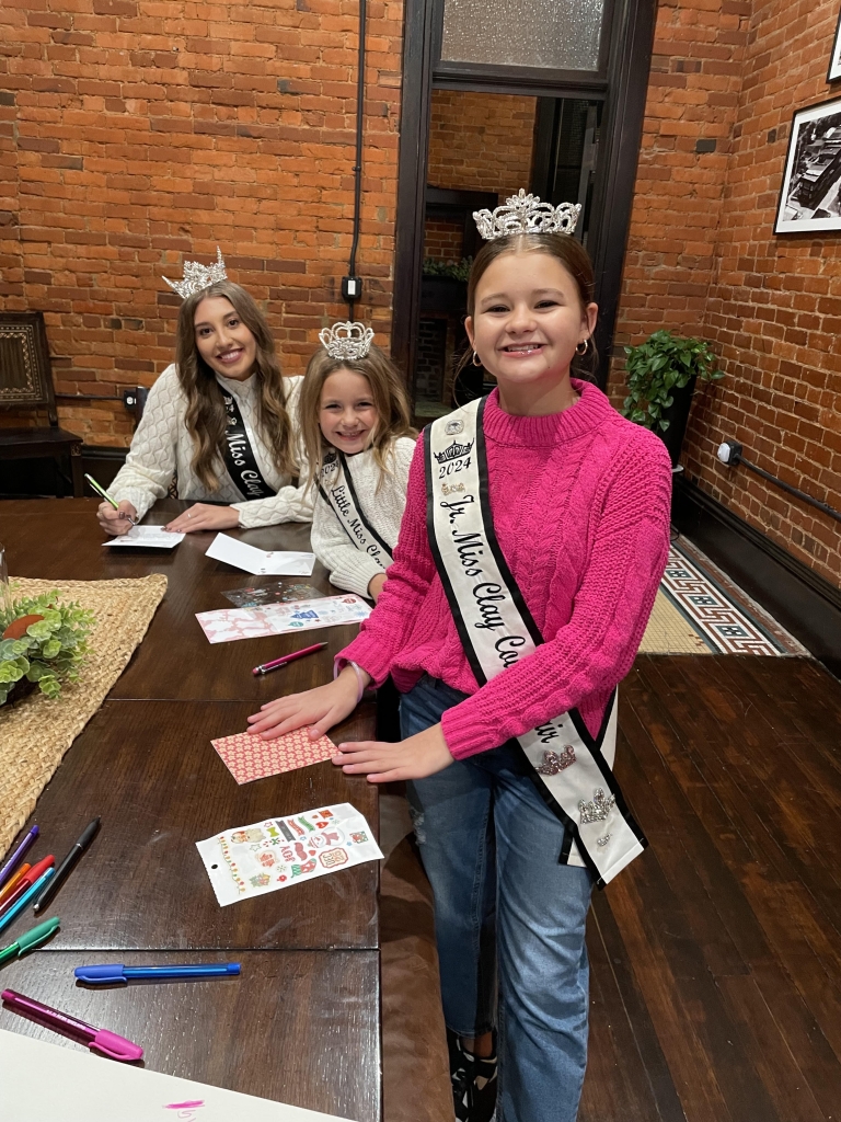 Clay County Fair Queens write cards at the Louisville Fall Festival to help Zink accomplish her goal.